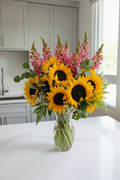 A vibrant bouquet of yellow sunflowers with dark brown centers and small white baby's breath, arranged in a clear glass vase on a white marble-topped side table.
