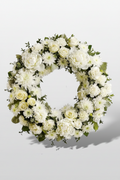 A circular floral wreath composed of white roses, white daisies, and green foliage, photographed against a light gray studio background.